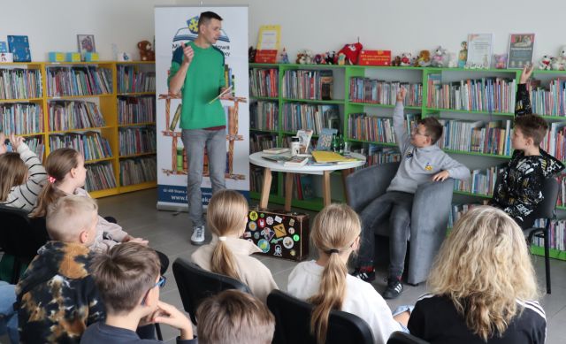 utor w zielonej bluzie stoi przed siedzącą na krzesłach młodzieżą, w tle regały z książkami, przy stoliku siedzi dwóch chłopców