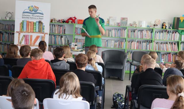 autor w zielonej bluzie stoi i czyta książkę przed siedzącą na krzesłach młodzieżą, w tle regały z książkami