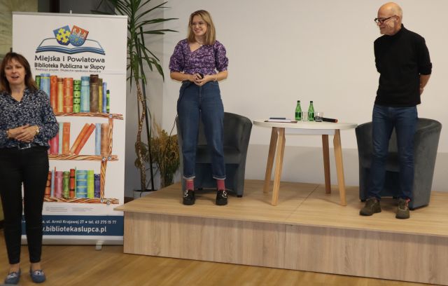 kobieta stoi przed sceną, na scenie stoi kobieta i mężczyzna, z ich lewej stronie baner biblioteki