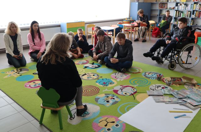 bibliotekarka siedzi na krzesełku, przed nią na dywanie siedzi młodzież
