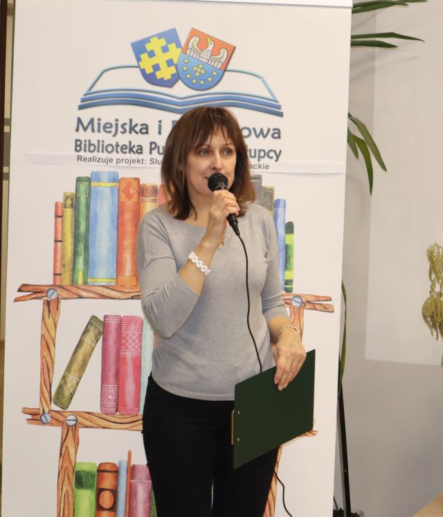 dyrektorka biblioteki z mikrofonem, w tle baner z nazwą biblioteki, logo i nazwą projektu