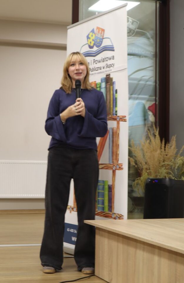 na zdjęciu stojąca dziewczyna z mikrofonem, w tle baner biblioteki