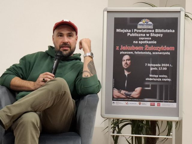 autor siedzący na fotelu z mikrofonem, w tle plakat z zapowiedzią jego spotkania