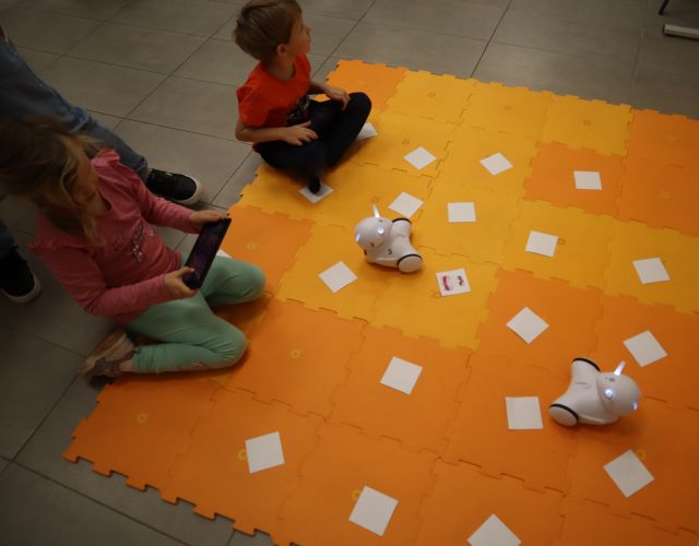 chłopiec i dziewczynka sterują robotami Photon przy użyciu tabletów, roboty na pomarańczowej macie, obok karteczki