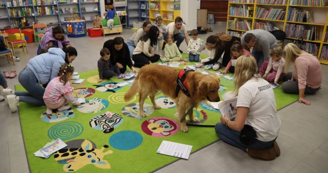 dzieci z rodzicami siedzą na kolorowym dywanie i układają obrazki, przed nimi siedzi opiekunka z psem