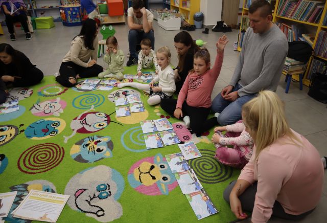 dzieci z rodzicami siedzą na kolorowym dywanie, przed nimi leżą obrazki