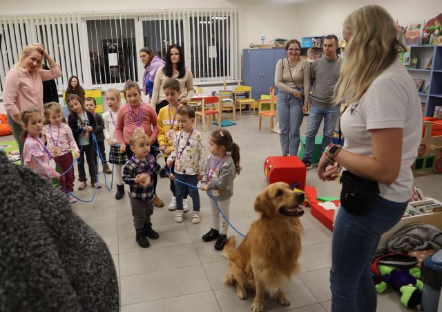 dzieci stoją w rządku trzymając psa na długiej smyczy, rodzice stoją obok, w tle regały