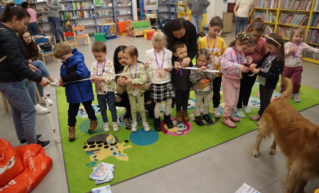 dzieci z rodzicami stoją w długim rządku i odklejają naklejki, w tle regały z książkami