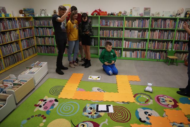 młodzież steruje rbotami przy użyciu tabletów, w tle regały z książkami