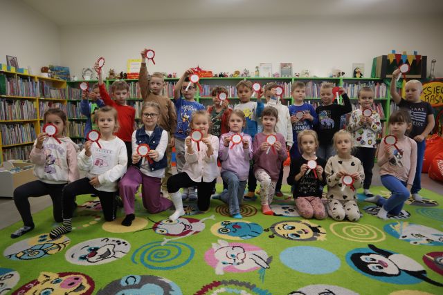 przedszkolaki pozujące do zdjęcia z wykonanymi przez siebie kotylionami, w tle regały z książkami