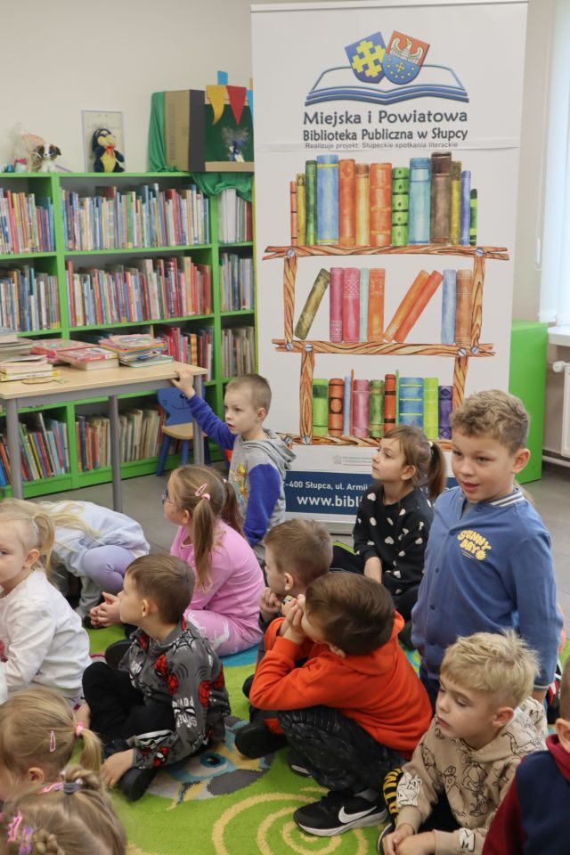 przedszkoalki siedzą na kolorowym dywanie, w tle baner biblioteki