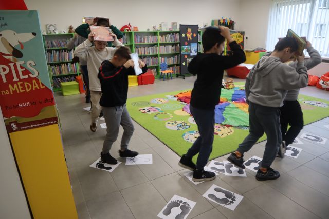 dzieci chodzą z książkami na głowie, w tle regały z książkami