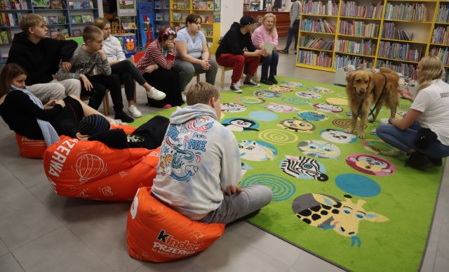 młodzież siedzi na krzesłach wokół kolorowego dywaniu, przed nimi kobieta z psem