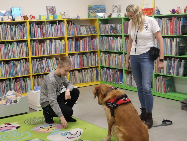 chłopiec kucający przed psem, obok psa stoi kobieta, w tle regały z książkami