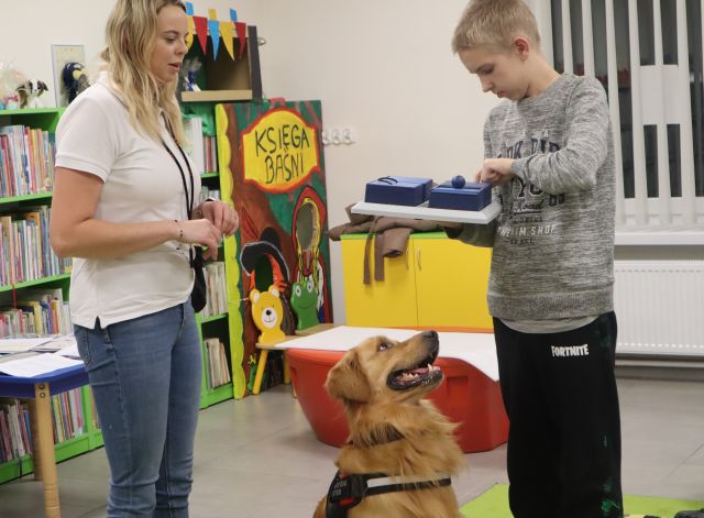 pomiędzy stojącą kobietą i chłopcem z niebieskimi pudełkami siedzi pies
