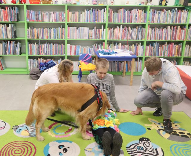 chłopic leży na dywanie, na nogach ma matę ze smakołykami, obok siedzi kobieta i kuca chłopiec, w tle regały z książkami