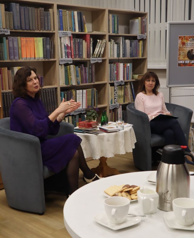 dwie kobiety siedzą na siwych fotelach, przy owalnym stoliku, w tle regały z książkami