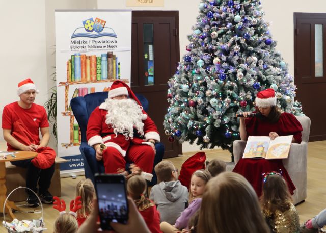 przy choince siedzi Mikołaj, Pani Mikołajowa z książką oraz elf