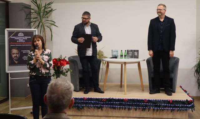 dyrektorka biblioteki z mikrofonem, na podwyższeniu stoi dwóch mężczyzn