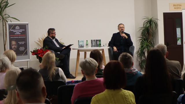 w tle przy okrągłym stole siedzi dwóch mężczyzn, przed nimi siedzą uczestnicy spotkania