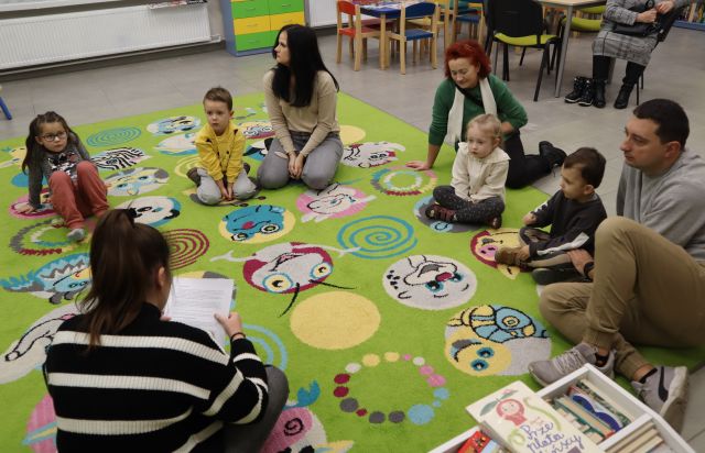dzieci z rodzicami i bibliotekarka siedzą na kolorowym dywanie