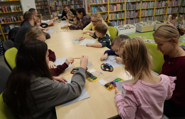 dzieci i rodzice siedzą przy połączonych stołach, na których leżą artykuły malarskie