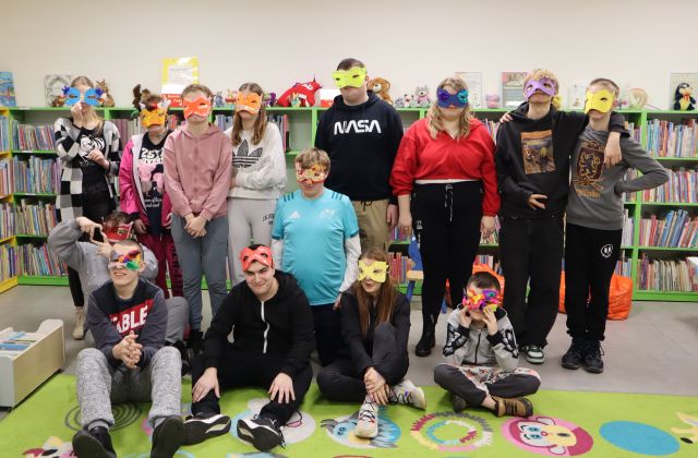 młodzież pozuje do zdjęcia w maskach karnawałowych, w tle regały z książkami