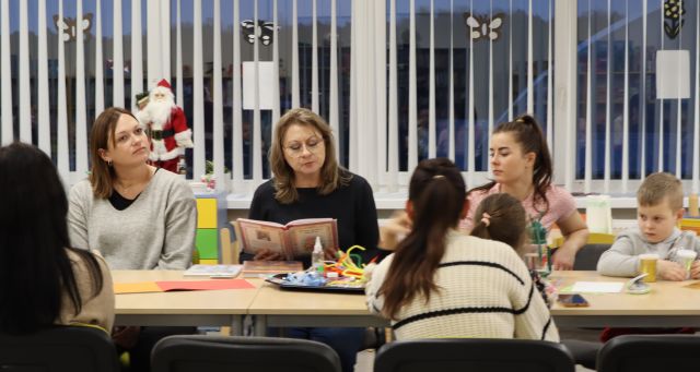 przy stole siedzą rodzice, dzieci i bibliotekarka z książką