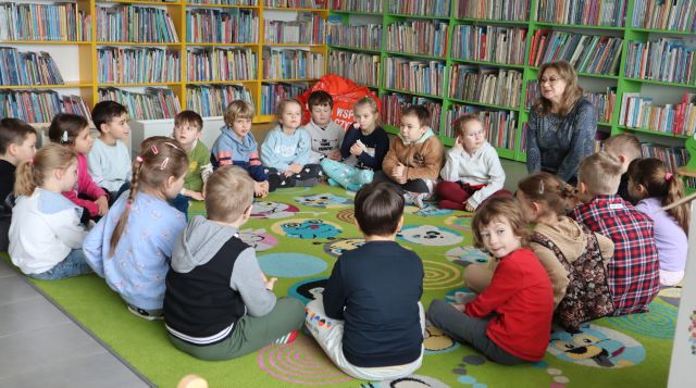 przedszkolaki z bibliotekarką siedzą w okręgu na kolorowym dywanie, w tle regały z książkami