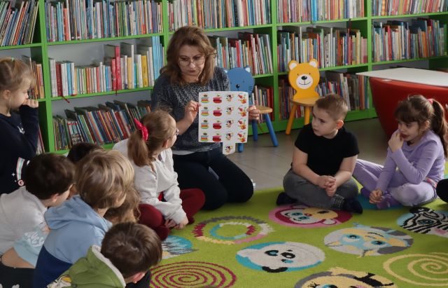 bibliotekarka pokazuje siedzącym wokół niej przedszkolakom kartkę z naklejkami, w tle regały z książkami