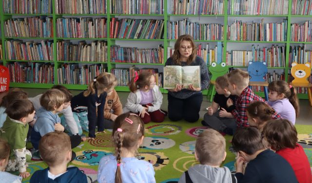 bibliotekarka siedzi i pokazuje siedzącym wokół niej przedszkolakom książkę, w tle regały z książkami