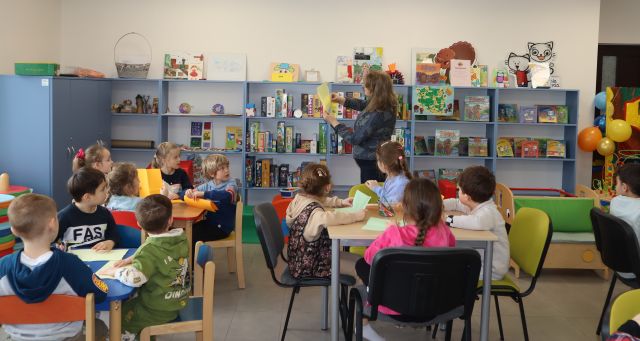 przed przedszkolakami siedzącymi przy stolikach stoi bibliotekarka, w tle regały