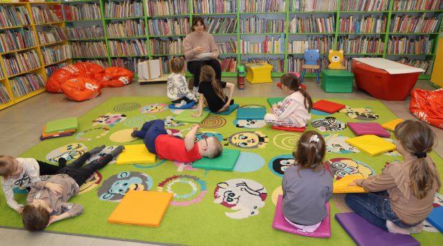 dzieci siedzą i leżą na kolorowym dywanie, przed nimi bibliotekarka, w tle regały z książkami