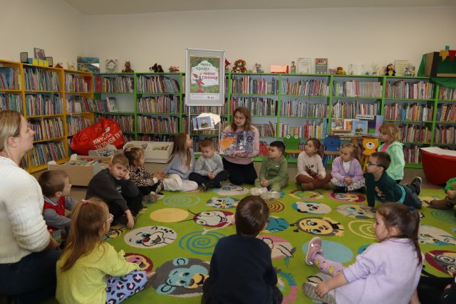 dzieci siedzą na kolorowym dywanie z bibliotekarką trzymającą książkę