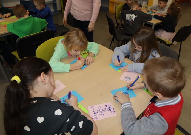 dzieci siedzą przy stole i malują na małych kartkach