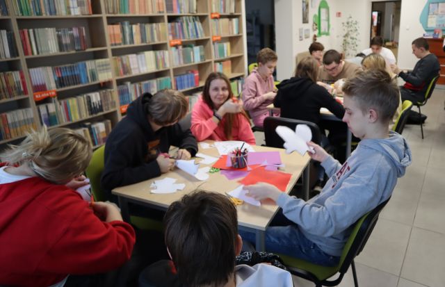 młodzoeż soiedzi przy stołach w bibliotece, na stołach materiały plastyczne