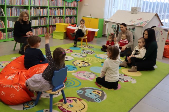 dzieci z rodzicami siedzą na kolorowym dywanie, przed nimi bibliotekarka siedzi na krzesełku, w tle regały z książkami