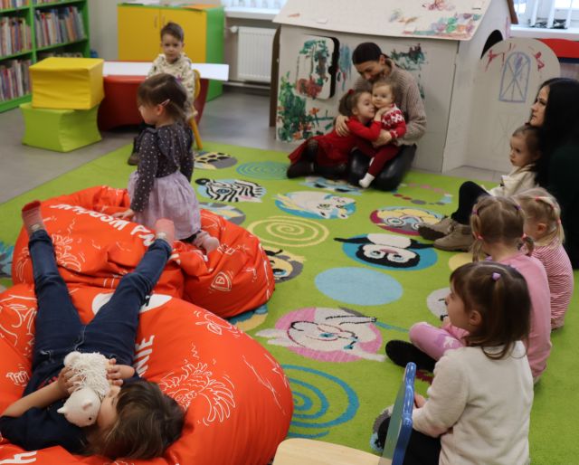 dzieci z rodzicami siedzą na kolorowym dywanie i leżą na kolorowych pufach,