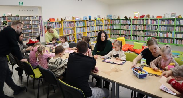 dzieci i rodzice siedzą przy stołach, na których leżą materiały papiernicze, w tle regały z książkami