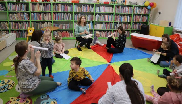 dzieci i rodzice siedą na chuście animacyjnej, przed nimi na krzesełku siedzi bibliotekarka z książką