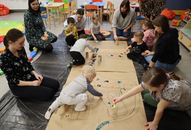 dzieci z rodzicami malują dużego dinozaura narysowanego na szarym papierze rozłożonego na podłodzea