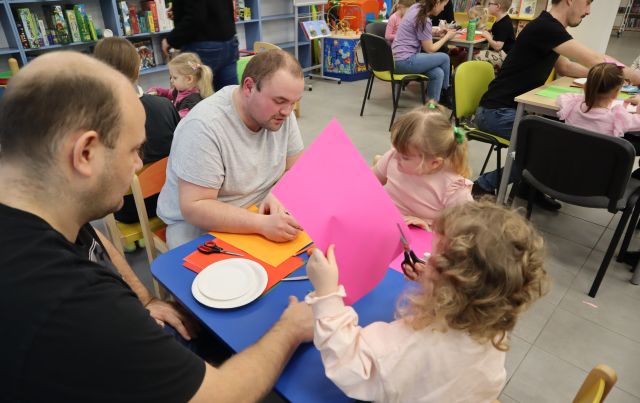 dzieci i rodzice siedzą przy stolikach i wycinają z kartek