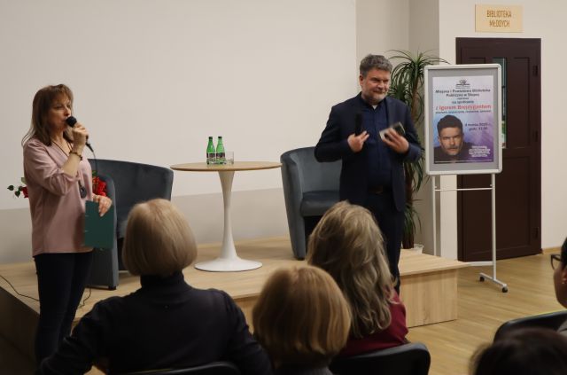 dyrektorka biblioteki i autor stoją z mikrofonami, przed nimi siedzą uczestnicy spotkania