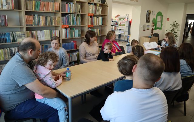 dzieci z rodzicami sieedzą przy połączonych stołach, w tle rgały z książkami