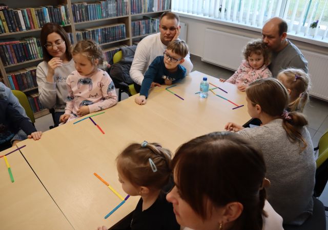 dzieci z rodzicami siedzą przy stołach, na których leżą koloowe patyczki ułożone w trójkąty, w tle regały z kiążkami