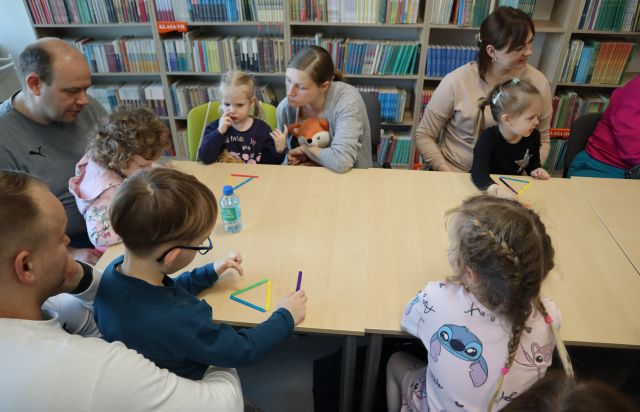 dzieci z rodzicami siedzą przy stołach, na których leżą koloowe patyczki ułożone w trójkąty, w tle regały z kiążkami