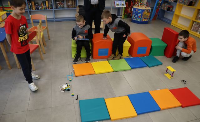 dwóch chłopców siedzi na kolorowych pufach, przed nimi na podłodze leżą kolorowe duże puzle i robot z klocków Lego