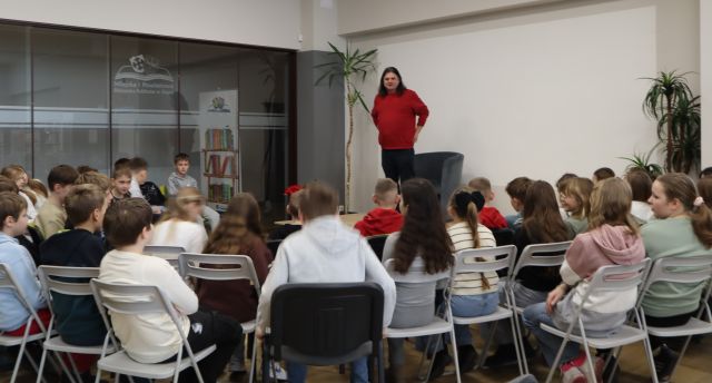 mężczyzna w czerwonym swetrze stoi na scenie, przed sceną siedzą na krzesłach czwartoklasiści z podstawówek