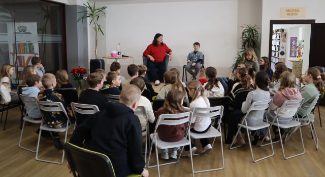 na scenie siedzi mężczyzna w czerwonym swetrze i chłopiec, przed nimi pod sceną siedzą dzieci