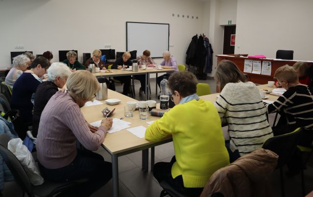 przy stołach siedzi blisko 20 seniorek i notuje na kartkach, w tle komputery i biurko